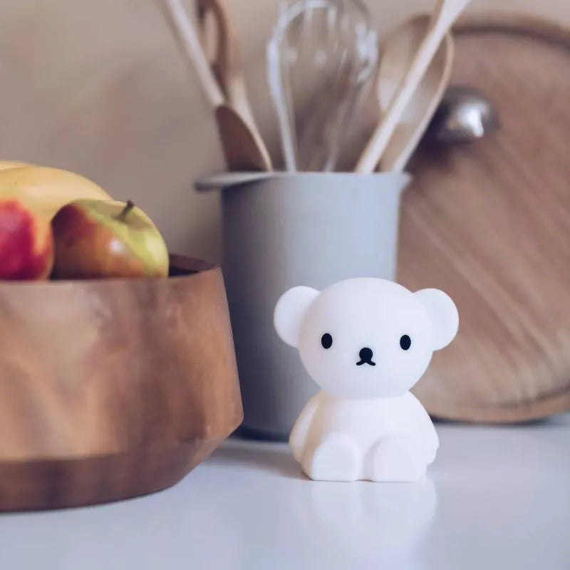 A Boris Bear Miffy Night Light, with its charming design, sits on a table beside a fruit bowl, appearing as if it's part of the delightful Miffy and Friends Light Series, nestled among kitchen utensils in a container.