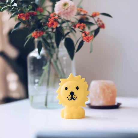 A Lion Miffy Night Light (15cm) from the Miffy brand, in a small yellow design, sits on a table with a glass vase of flowers and a candle in the background.