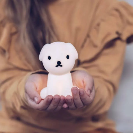 The individual, dressed in a beige sweater, gently holds a Snuffy Dog Miffy Night Light (15cm) in their hands—a delightful dog-shaped lamp that emits a gentle glow. Constructed from BPA-free silicone, this charming piece by Miffy infuses any space with a whimsical touch.