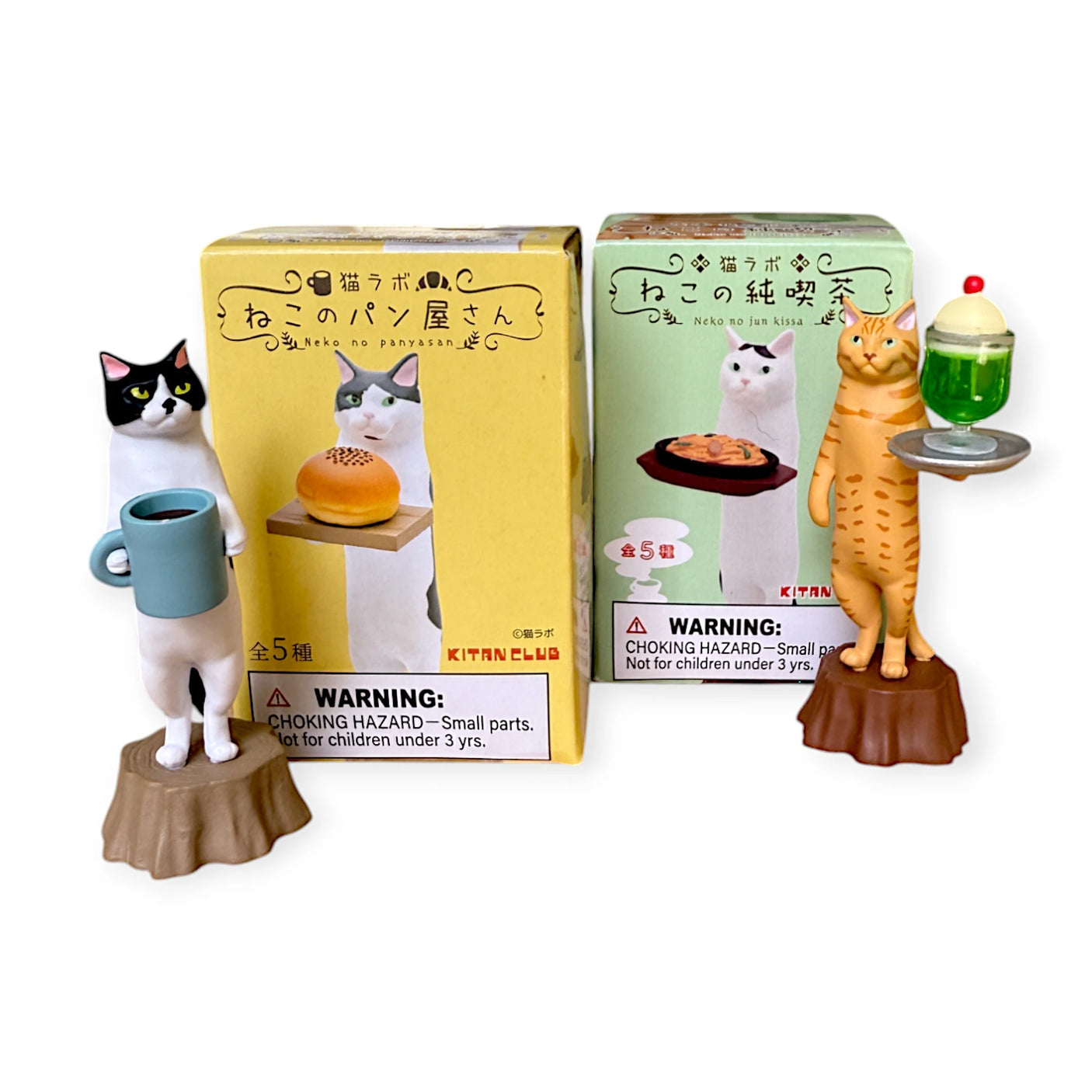 Two Cat Cafe figurines from Kitan Club stand upright: one as a waiter with coffee and a bread box, the other with dessert and a green box. The Cat Cafe Waiter Blind Box packaging includes warnings about small parts not suitable for children under 3 years old.