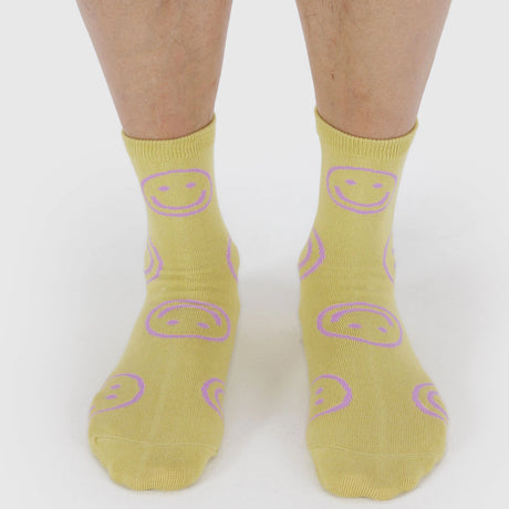 Feet wearing Baggu crew socks in warm straw yellow with lavender smiley faces, front view, product image one