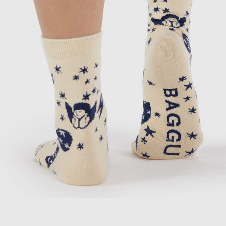 Someone wears Baggu Crew Socks - Cherub Bows by Baggu, featuring a beige and blue pattern with bows, "BAGGU" on the heel, made from a soft bamboo rayon blend, standing against a light background.