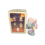 A toy figurine with pink hair and a magnifying glass stands beside a Lucky EMMA - Secret Forest of Dim Lights Series Blind Box, offering a glimpse into the whimsical adventures from the Lucky Emma brand.