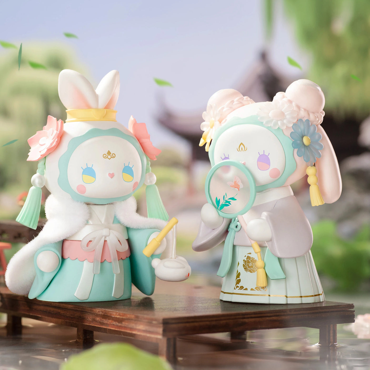 Two whimsical figurines from the Lucky EMMA - Secret Forest of Dim Lights Series Blind Box, dressed in traditional Asian attire with rabbit-like features, stand gracefully on a wooden platform. They are encircled by lush greenery and captivating architecture, capturing the mystery and charm associated with Lucky Emma's enchanting collection.