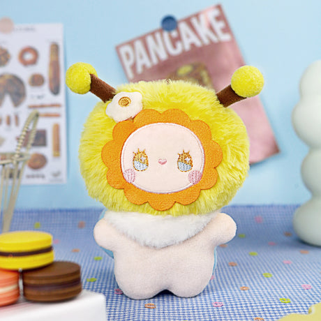 The Emma Fuzzy Family Keychain Blind Box by Lucky Emma features a cute plush charm with a yellow lion-like mane, small horns, and flower detail, shown on a blue dotted surface near macarons and a "PANCAKE" sign.