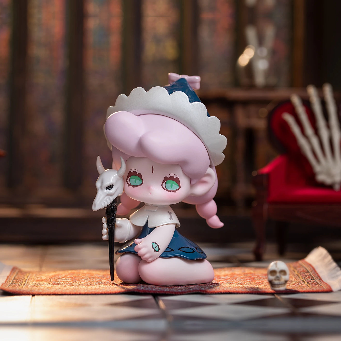 A petite figure with pink hair, dressed in a maid outfit and holding a skull staff, sits on a patterned rug. Surrounded by skeletal decorations and resting against the red chair, the aura hints at Lucky Emma's Killa Treasure Pavilion Blind Box mysteries eager to be unveiled.