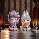 Two playful "Lucky EMMA - Killa Treasure Pavilion Blind Box" figurines, featuring pastel hues, are placed on a checkered floor, encircled by candles in the softly lit atmosphere of the Killa Treasure Pavilion.