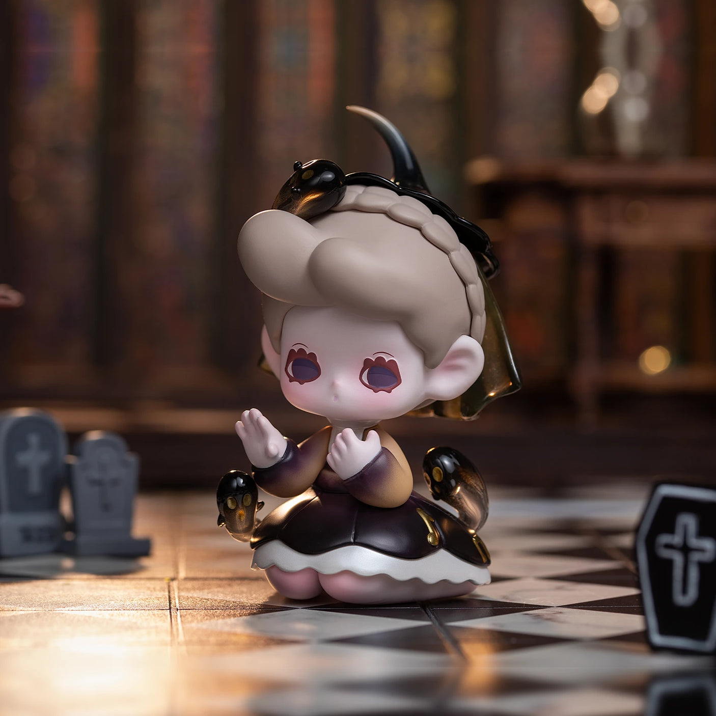 A charming figurine with heart-shaped eyes and bat wings is seated on a checkered floor, encircled by petite tombstones in a room featuring stained glass windows. This mesmerizing piece is from the Lucky EMMA - Killa Treasure Pavilion Blind Box collection, providing an enticing peek into Secret Treasures that inspire the imagination.