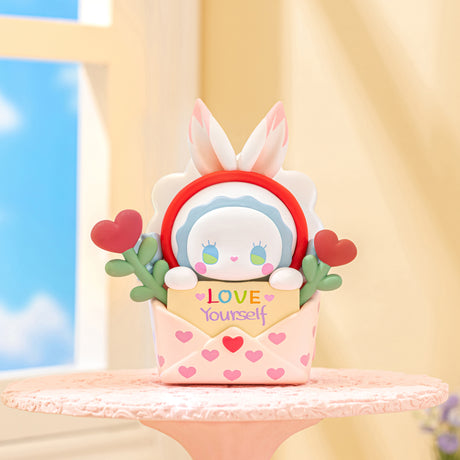 Emma Secret Forest Love Dopamine figure with bunny ears holding a "Love Yourself" card inside a heart-patterned envelope on table