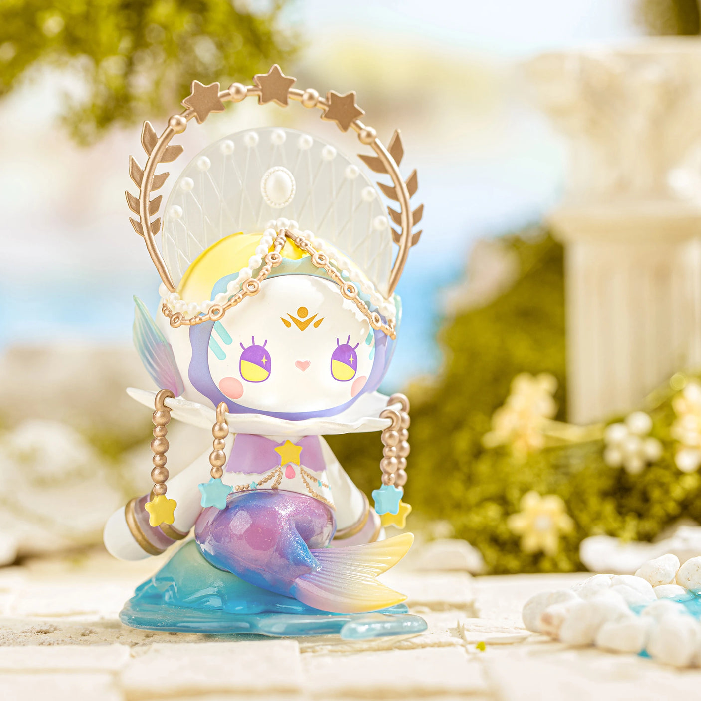 A captivating figurine from the Lucky EMMA - Secret Forest River of Time Blind Box series, this colorful piece is adorned with a celestial crown and holds a mermaid tail. Situated outdoors, the figurine is surrounded by lush greenery and blurred stones, capturing the enchanting mystery characteristic of Lucky Emma creations in a blind box surprise.