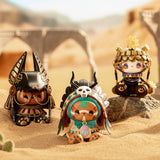 Three small, vibrant figurines from the Lucky Emma's River of Time Blind Box collection, each adorned in ancient-themed costumes, are displayed on sand against a desert backdrop with stone arches.