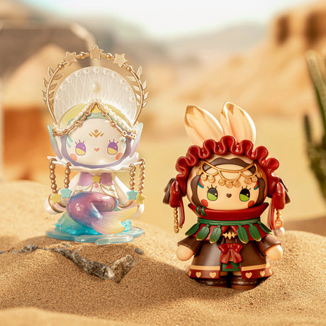 Two vibrant figures from the Lucky EMMA - Secret Forest River of Time Blind Box collection stand on a sandy terrain. One displays a fish and seashell theme, while the other showcases gold and red accessories with feathery accents. Set against a blurred desert backdrop, these figures evoke a mysterious air reminiscent of a blind box surprise from the Lucky Emma brand.