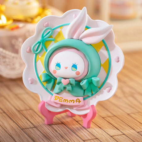 A charming, hand-painted figurine featuring bunny ears and a green outfit stands on pink legs, reminiscent of the products from Lucky Emma. The front displays the name "Yemma." Ideal for a Secret Forest Birthday Party backdrop, it is complemented by a softly blurred background illuminated with warm light.