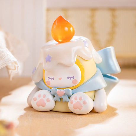 The charming Lucky EMMA - Secret Forest Birthday Party Blind Box figure from the Lucky Emma brand showcases a character with frosting-like hair, a glowing flame on top, a blue ribbon, and paw-shaped feet. It evokes the whimsical style of the Emma Figures collection and looks delightful when placed on a wooden surface.