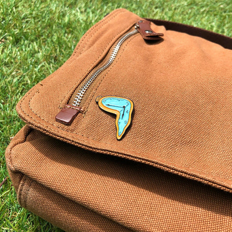 Dali Persistence of Memory enamel pin with melting clock design on brown canvas bag flap outdoors, gallery image 1.webp