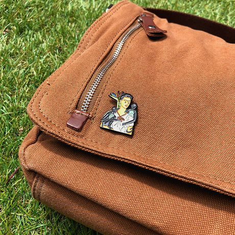 Enamel pin showing Frida Kahlo with two monkeys on brown bag flap outdoors, gallery image 1, file enamel-pin_Frida_Khalo_Monkeys.webp