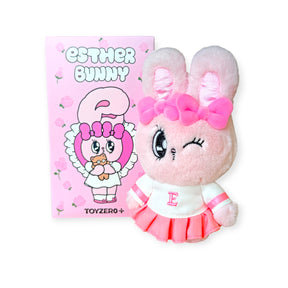 Pink Esther Bunny plush keychain with bow and skirt, winking, next to illustrated pink packaging box with floral design