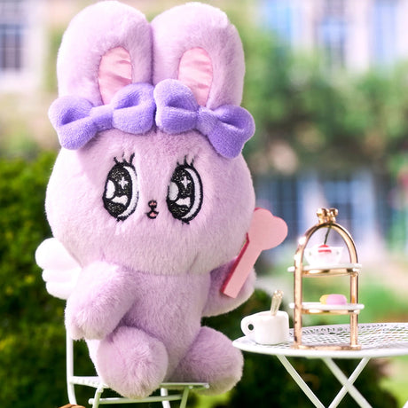 Lilac Esther Bunny plush keychain with purple bow holding pink heart seated at white table with tea set outdoors
