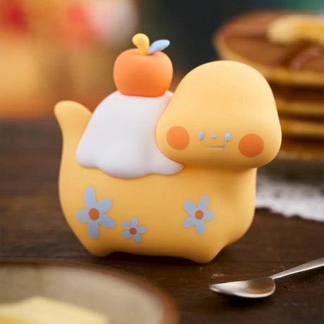 Soft yellow Food Dinosaur Fuji Yama Vol. 2 figure with white frosting, peach fruit, and blue floral details on wooden table close-up