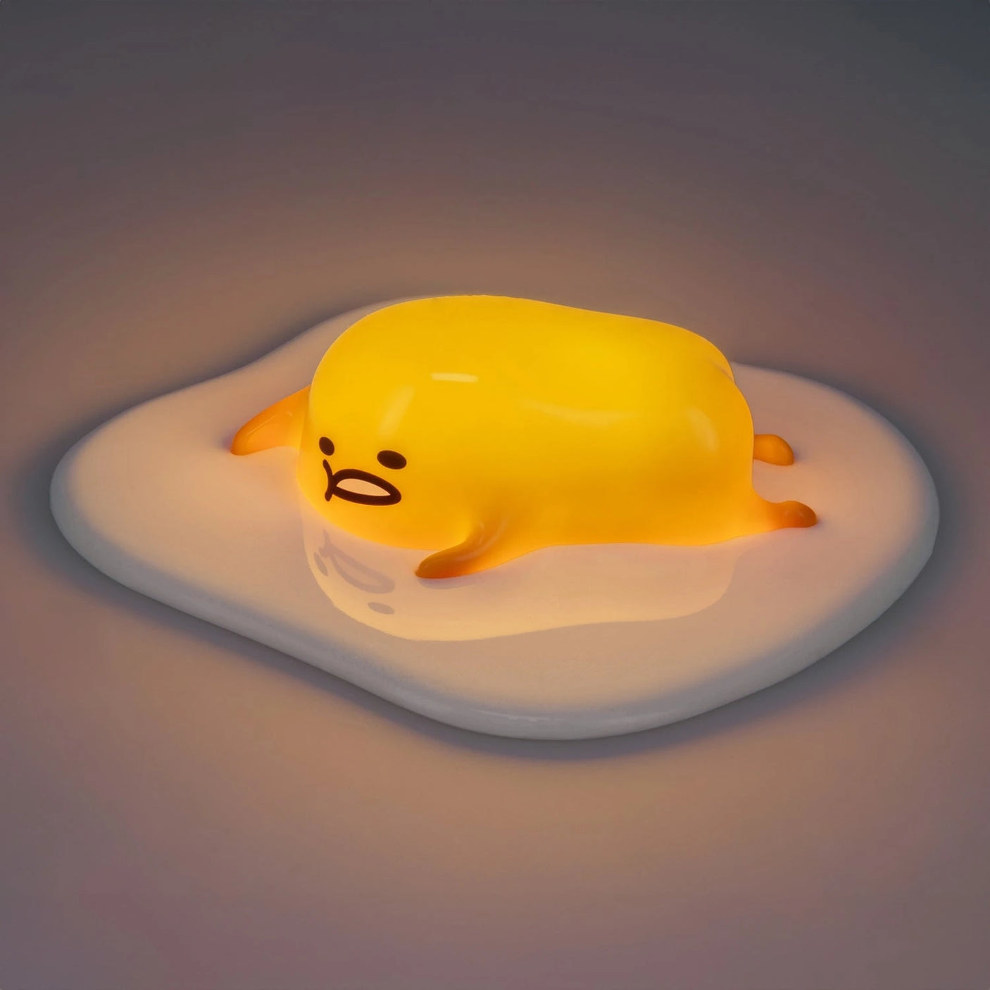 A yellow, blob-like character with a lazy expression rests on a white surface resembling an egg white, similar to the Gudetama 3D Squishy Silicone Night Light by Gudetama.