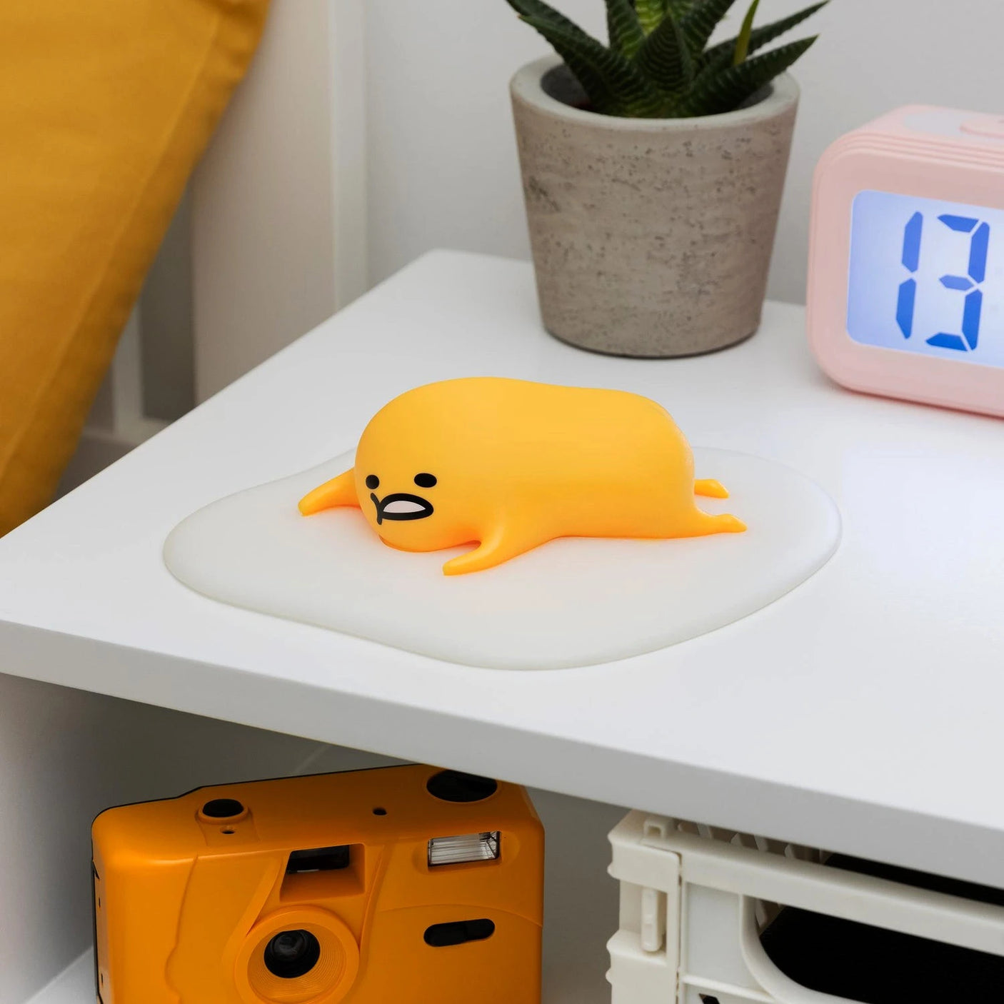 A squishy silicone toy shaped like Gudetama, known as the Gudetama 3D Squishy Silicone Night Light by Gudetama, lies on a white surface alongside a plant, clock, and orange camera.