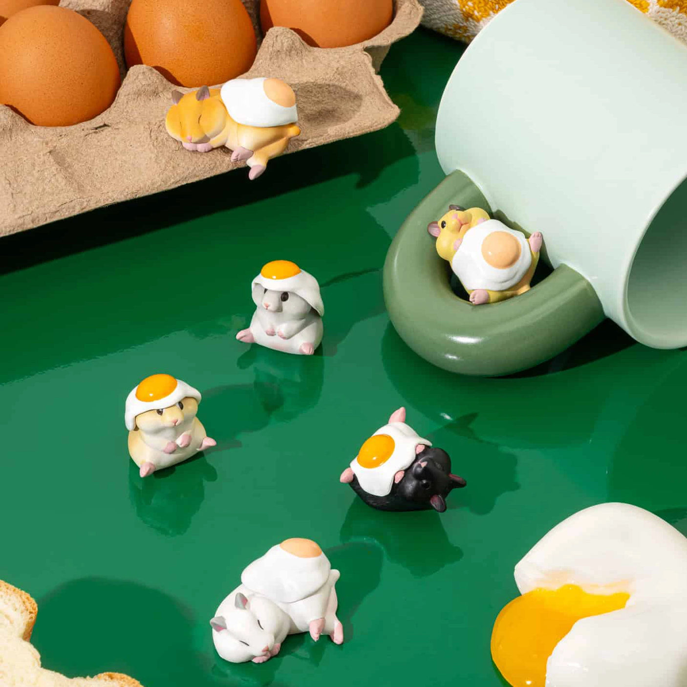 The Hamster ‘N Egg Blind Box mini figures by Kitan Club, featuring small animal figurines with egg decorations, are displayed on a green surface next to a tipped container and brown eggs in a carton.