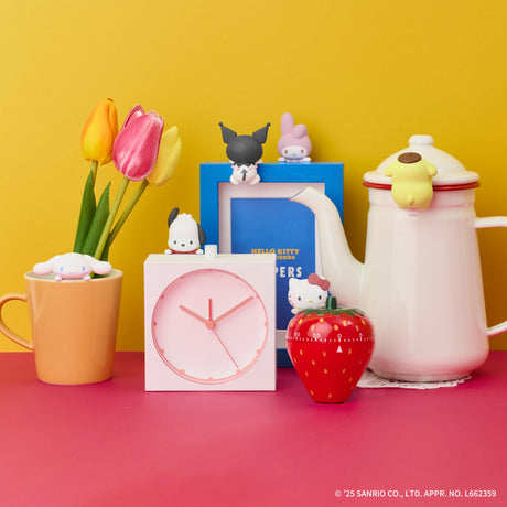 Pastel character figurines on pink clock, orange cup with tulips, blue book, red strawberry container, and white teapot on pink surface