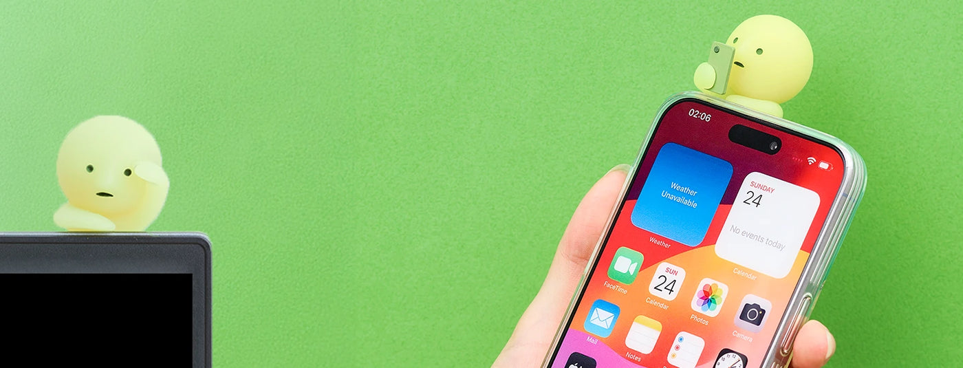 Smartphone with colorful home screen held against a green background with a small, round yellow figure on top. Another figure is on a computer monitor in the background.