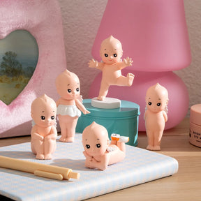Five small baby dolls with varied poses arranged on tabletop near pink lamp and decorative items