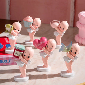 The Smoko Kewpie Holding Things Blind Box features six small angelic vinyl figures, each holding a bird, cupcake, teapot, jam jar, heart, or butterfly. Displayed on white stands against a pink backdrop.