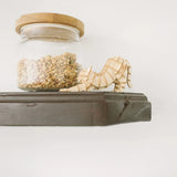 A jar of granola and the "Cat Stretching Wooden Puzzle" by papernthought, crafted from FSC Certified plywood, rest on a dark wooden shelf against a white wall.