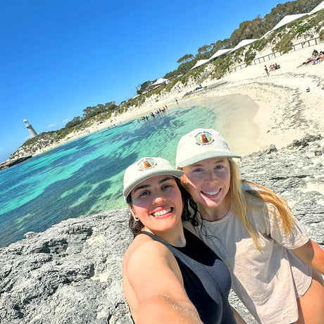 Two women wearing off-white Lies Collective hats with smiling quokka embroidery posing on rocky beach near turquoise water and lighthouse