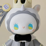 A close-up of the Lucky EMMA - Secret Forest Wedding toy figure from Lucky Emma, showcasing its round face, blue and yellow eyes, gray mane, small horn, crown, and black bow tie.