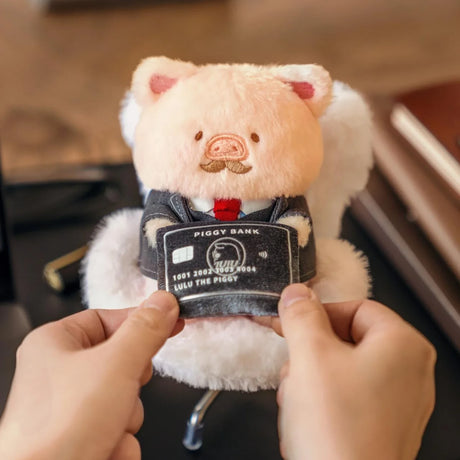 Plush piggy keychain held in hands, wearing a dark suit with piggy bank label.