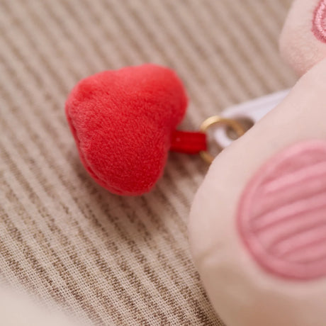 Close-up of plush red heart charm attached to soft pink LuLu the Piggy pouch bag accessory on fabric surface