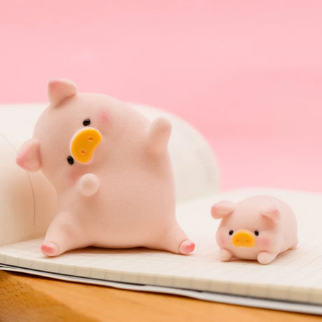 Two soft-textured LuLu the Piggy Original Series 2 figures posed on an open notebook with pink background
