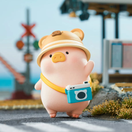 LuLu the Piggy figure wearing a beige hat and carrying a blue camera toy standing on a road with blurred outdoor background
