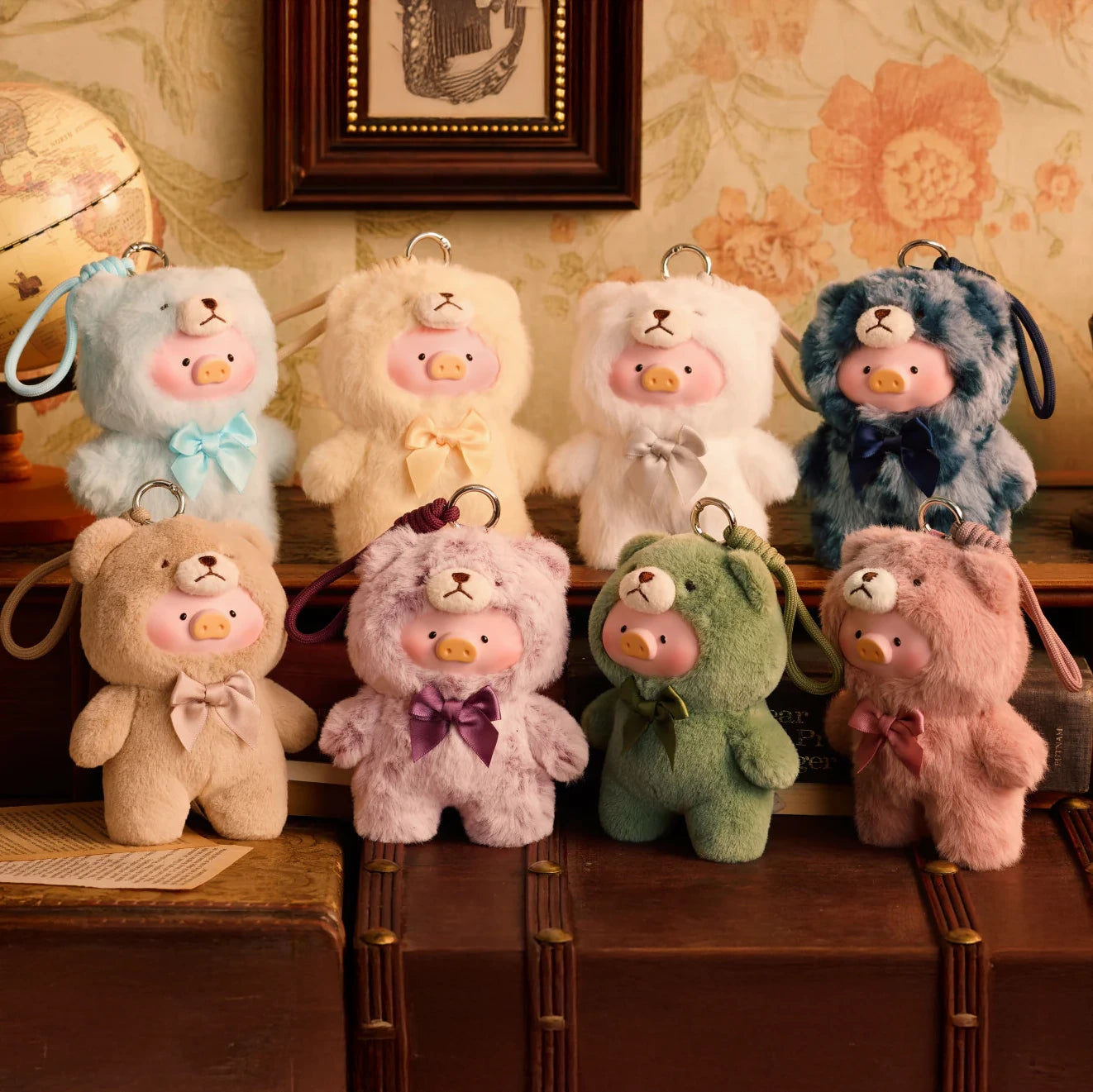 Seven Teddy LuLu the Piggy Plush Pal – Blind Box keychains by LuLu The Piggy, in assorted colors with bows, are displayed on books with floral wallpaper—ideal for Blind Box Plush collectors.