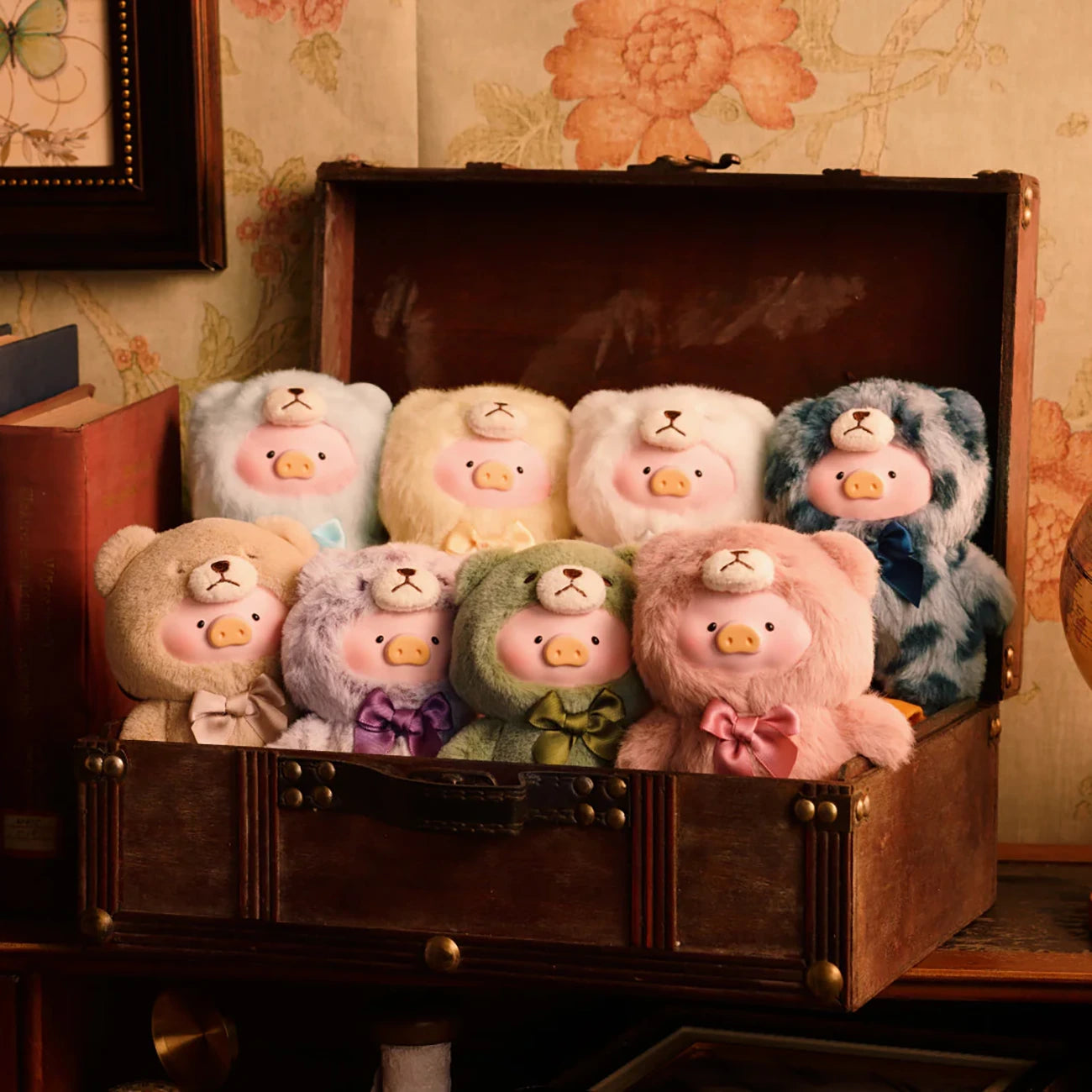 A wooden chest holds nine LuLu The Piggy "Teddy LuLu the Piggy Plush Pal - Blind Box" toys, each with a pastel bow, displayed on a table against floral-patterned wallpaper.