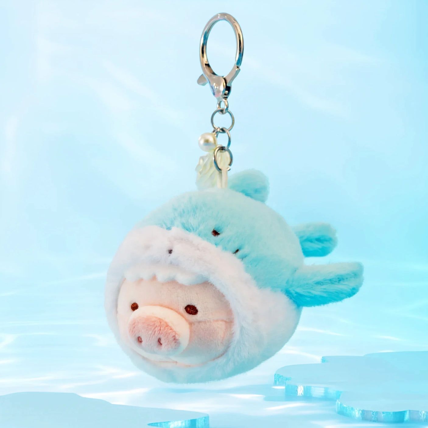The LuLu The Piggy - Vibrating Shark Keychain features LuLu the Piggy in a light blue dolphin costume, hanging against a soft blue background with ice accents—a cute accessory for fans of adorable Shark Plush items.