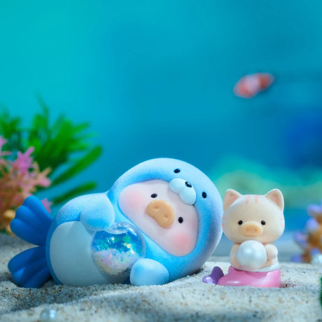 A blue seal-like character with a shiny ball lounges on sand next to a LuLu The Piggy - Ocean Blind Box figurine holding a pearl, both from the LuLu The Piggy brand, surrounded by aquatic plants and blurred fish for ocean adventure vibes.