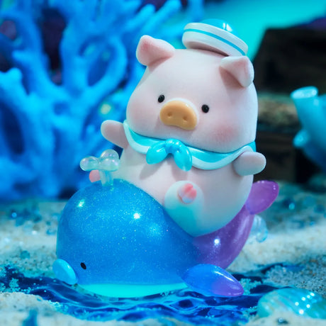 A LuLu The Piggy - Ocean Blind Box toy from LuLu The Piggy sits in a sailor hat and scarf atop a sparkling blue and purple whale, with blue coral and water decorations for an ocean-themed display.