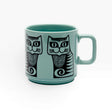 Teal ceramic mug with two black-and-gray cat faces and striped tails