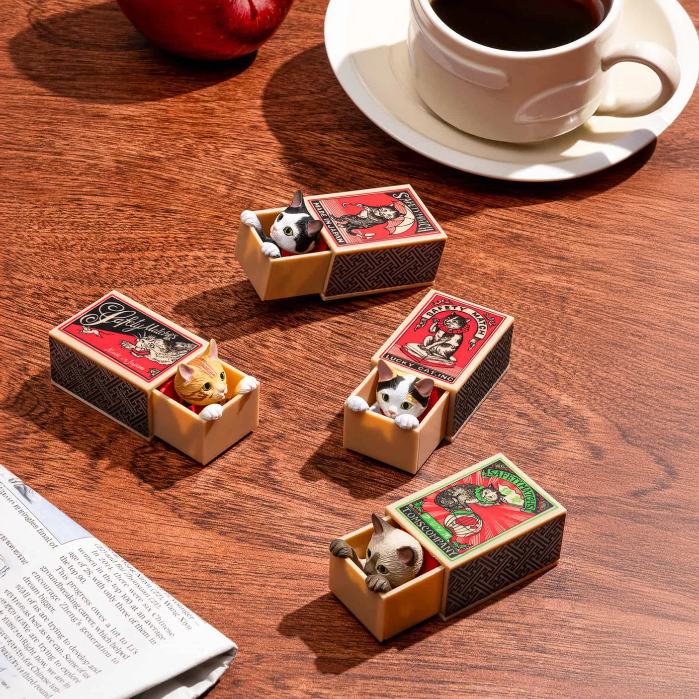 Four matchboxes, each containing a Cat in a Matchbox Blind Box from Kitan Club, sit on a wooden table beside a cup of coffee, an apple, and a newspaper.