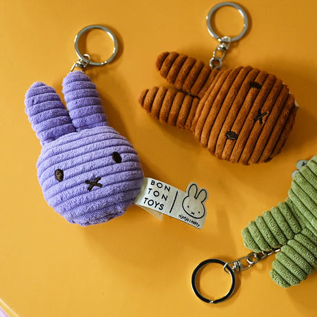 Three Miffy Flat Corduroy Keychains in purple, brown, and green are arranged on a yellow surface. The tags read "Bon Ton Toys," adding texture to this adorable collection featuring the Miffy brand.