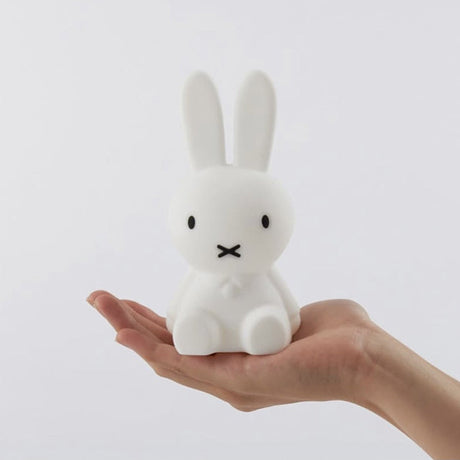 A hand holds a small, white Miffy Bunny Night Light (15cm) with black eyes and a simple design, crafted from BPA-free silicone by the brand Miffy, against a plain background.