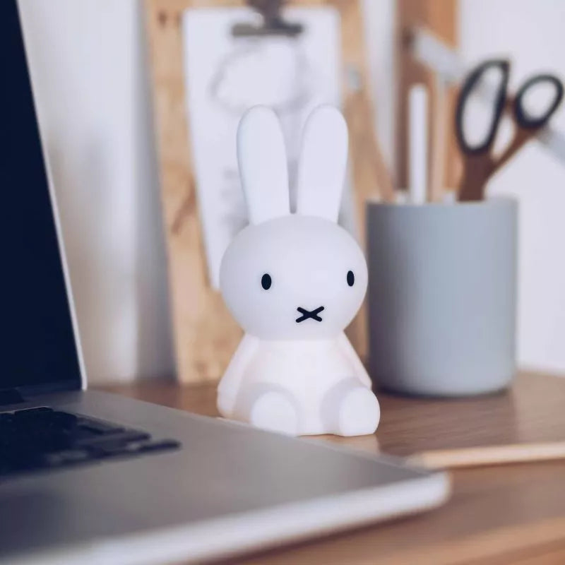 A Miffy Bunny Night Light (15cm) from the Miffy brand, crafted from BPA-free silicone, sits next to a laptop on a wooden desk, with stationery scattered in the background.