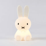 The Miffy Bunny Night Light, a 15cm lamp from the Miffy brand, features a glowing white design with long ears and minimal facial features. Made from BPA-free silicone for safety, it serves as a comforting nightlight against a plain backdrop.