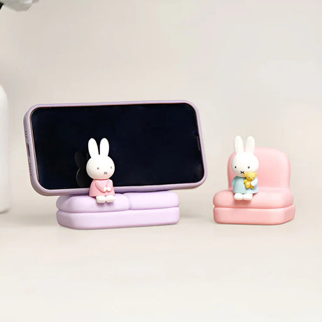 Two mini Miffy and Dan figures seated on pastel purple and pink sofa stands, one holding phone and other holding teddy bear