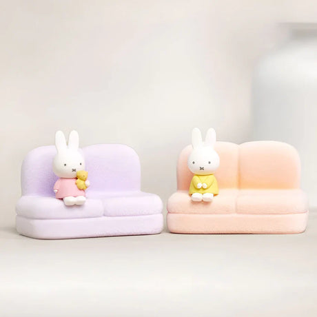 Two mini Miffy figures wearing pastel outfits seated on soft lavender and peach sofa stands against plain background